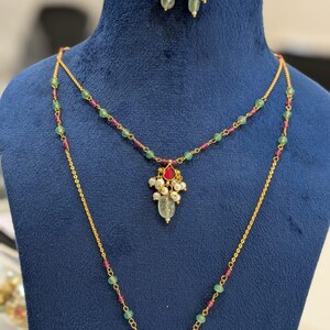 May include: A gold-colored necklace and earring set displayed on a blue mannequin. The necklace features a double strand design with green and red beads, and a pendant with a red gem, pearls, and a green teardrop-shaped stone. The earrings have a similar design.