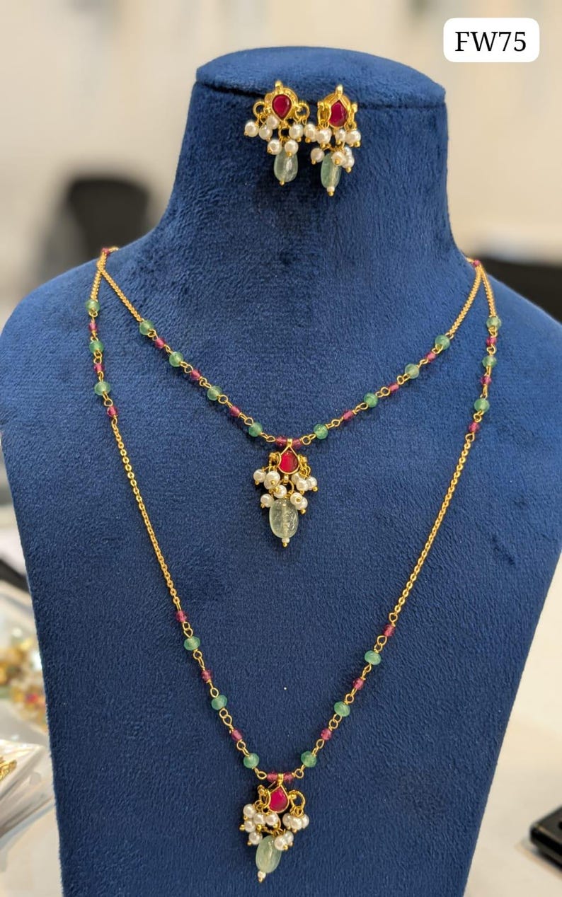 May include: A gold-colored necklace and earring set displayed on a blue mannequin. The necklace features a double strand design with green and red beads, and a pendant with a red gem, pearls, and a green teardrop-shaped stone. The earrings have a similar design.