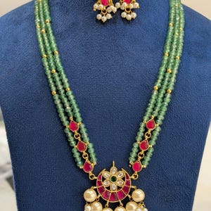May include: A three-strand green beaded necklace with a pendant and matching earrings. The pendant features a red and gold design with white pearls. The earrings have a similar design with red stones and white pearls. The necklace and earrings are displayed on a blue surface. The text "FW175" is visible.