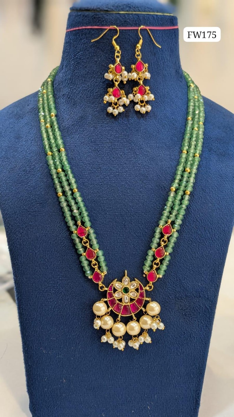 May include: A three-strand green beaded necklace with a pendant and matching earrings. The pendant features a red and gold design with white pearls. The earrings have a similar design with red stones and white pearls. The necklace and earrings are displayed on a blue surface. The text "FW175" is visible.