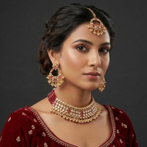 May include: A gold-toned jewelry set featuring a choker necklace, dangling earrings, and a maang tikka. Each piece is adorned with small red stones, clear crystals, and tiny gold beads. The set is worn with a deep red velvet garment with embroidery.