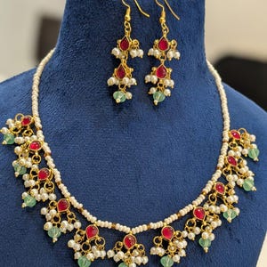 May include: A gold-toned necklace and earring set with red teardrop-shaped stones, white pearls, and green beads. The necklace and earrings are displayed on a blue velvet bust. The text "FW100" is in the upper right corner.