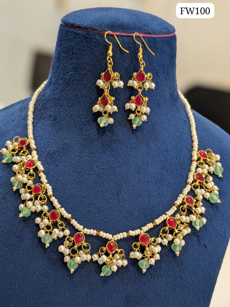 May include: A gold-toned necklace and earring set with red teardrop-shaped stones, white pearls, and green beads. The necklace and earrings are displayed on a blue velvet bust. The text "FW100" is in the upper right corner.