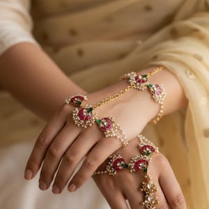 Op de afbeelding: Goudkleurige handjuwelen met rode en groene accenten en kleine witte parels. Het ontwerp omvat een ring en een armband die met elkaar verbonden zijn door een delicate ketting. De sieraden worden op de hand gedragen.
