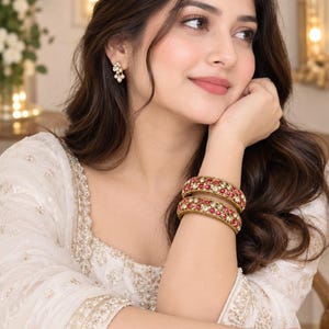 May include: A pair of gold-toned bangles featuring circular red stones and small white pearls, paired with matching cluster earrings. The jewellery set is worn with a white garment featuring intricate gold embroidery and delicate beadwork details.