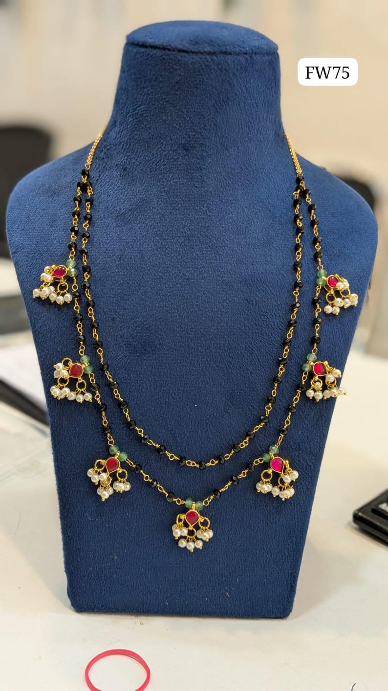 May include: A gold-toned necklace with two strands of black beads. The necklace features red and white accents, with small pearls and green beads. The text "FW75" is visible in the upper right corner.