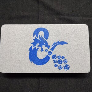 Peut inclure: Un boîtier rectangulaire en métal argenté arborant une impression graphique bleue représentant un symbole esperluette en forme de dragon, avec un groupe de diverses formes de dés polyédriques sortant de la gueule du dragon sur le couvercle.