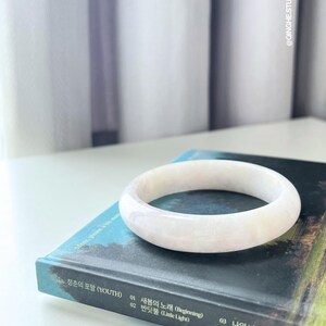 Könnte beinhalten: Ein hellrosa Jade-Armreif liegt auf einem Buch mit koreanischem Text. Das Armband ist glatt und kreisförmig. Das Buch hat einen blauen Einband mit koreanischem Text und den Wörtern "YOUTH" und "Time Machine". Der Hintergrund ist eine weiße Oberfläche und ein weißer Vorhang.
