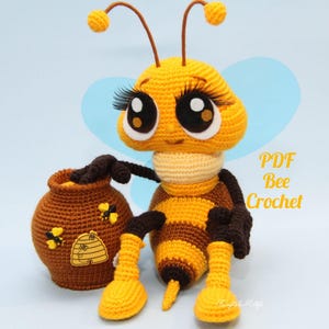 May include: A handmade crochet bee with large eyes, eyelashes, and antennae, next to a brown honey pot. The bee is yellow and brown with a cream-colored neck. The honey pot has a beehive and bee applique. The text "PDF Bee Crochet" is visible.
