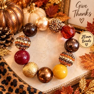 May include: A circular arrangement of 20mm beads in various finishes including metallic gold, deep brown, burgundy, and amber. Some beads feature a smooth glossy surface, while others are covered in sparkling crystals or a textured glitter finish.