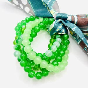 May include: A set of three green beaded bracelets in varying shades of green, tied with a patterned teal and brown scarf. The beads are round and appear to be made of a smooth, glossy material.