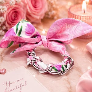 May include: A hair scrunchie featuring a silver-toned metal chain link band interwoven with a pink and white fabric ribbon. The ribbon is tied into a bow at the top and displays a floral pattern with pink flowers and green leaves on one side.