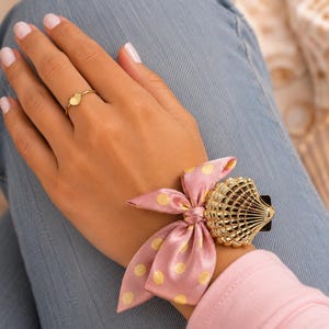 May include: A gold-toned metal ring featuring a small heart-shaped charm and a wrist accessory consisting of a metallic gold seashell-shaped piece attached to a pink fabric ribbon with a yellow polka dot pattern tied in a bow.