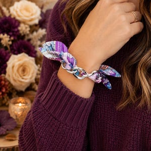 May include: A bracelet featuring a silver-toned metal chain link design intertwined with a patterned fabric ribbon. The fabric displays a vibrant mix of purple, blue, and white geometric and abstract shapes, tied in a knot around the metallic links.