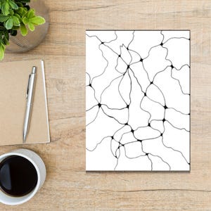 Könnte beinhalten: Ein weißer Druck mit einem schwarzen abstrakten Design aus miteinander verbundenen Linien und Knoten. Das Kunstwerk wird auf einem Holztisch präsentiert, zusammen mit einem Notizblock, einem Stift, einer Kaffeetasse und einer kleinen Pflanze in einem silbernen Topf. Die Gesamtästhetik ist minimalistisch.