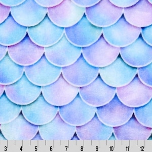 May include: A watercolor fabric featuring a mermaid scale pattern. The design showcases overlapping, rounded shapes in shades of blue, purple, and pink. A ruler is at the bottom, with markings in inches.