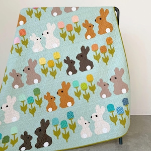May include: A light blue quilt featuring a repeating pattern of small and large rabbits in shades of white, brown, and dark gray, interspersed with stylized flowers in yellow, orange, pink, and teal on green stems with a dark green border.