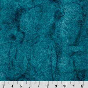 May include: Close-up of a teal-colored fabric with a textured, plush appearance. The fabric has a soft, fur-like texture, with visible ridges and valleys. The image includes a ruler along the bottom edge, with measurements in inches.