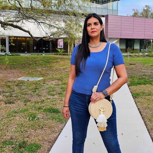 Puede incluir: Una mujer con un top azul y vaqueros oscuros, lleva un bolso redondo tejido con una estrella de mar y borlas. Lleva un collar turquesa y un reloj. El fondo muestra un edificio y un &aacute;rbol.