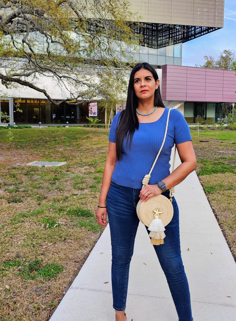 Puede incluir: Una mujer con un top azul y vaqueros oscuros, lleva un bolso redondo tejido con una estrella de mar y borlas. Lleva un collar turquesa y un reloj. El fondo muestra un edificio y un &aacute;rbol.