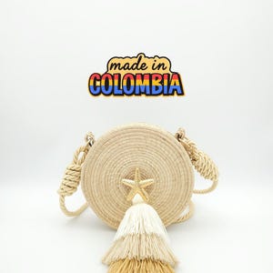 Puede incluir: Bolso redondo de paja tejida con borla crema y detalle de estrella de mar dorada. El bolso tiene asas de cuerda y el texto "made in COLOMBIA" en un dise&ntilde;o colorido.