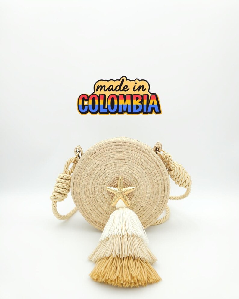 Puede incluir: Bolso redondo de paja tejida con borla crema y detalle de estrella de mar dorada. El bolso tiene asas de cuerda y el texto "made in COLOMBIA" en un dise&ntilde;o colorido.