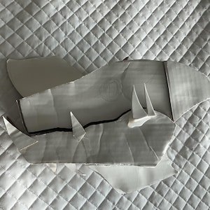 Puede incluir: Un conjunto de recortes de papel blanco, posiblemente patrones o plantillas, dispuestos sobre una superficie acolchada blanca. Los recortes tienen ángulos agudos y parecen ser parte de un diseño más grande. La iluminación crea sombras, agregando profundidad a la composición.
