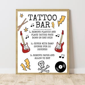 May include: A rectangular sign with a wooden frame featuring the text TATTOO BAR and instructions for application. The design includes illustrations of two red electric guitars, a black vinyl record, a skull with crossbones, lightning bolts, and musical notes.