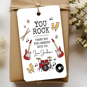 May include: A rectangular white gift tag featuring black text that reads YOU ROCK THANK YOU FOR JAMMING WITH US! and Love Jackson. The design includes illustrations of two red electric guitars, a red drum set with the number 3, lightning bolts, and music notes.