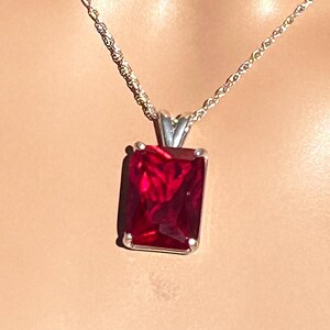 May include: A silver chain necklace with a red gemstone pendant. The gemstone is a pyrope garnet, set in an emerald style. The text on the image reads "Pyrope Garnet Red Sparkle Example of Dark Setting Emerald Style".