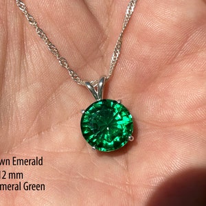 May include: A silver necklace with a dark emerald green, lab-grown, 12 mm round pendant. The pendant is a vibrant, faceted gemstone. The necklace is displayed on a hand.