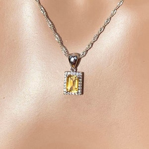 May include: A rectangular yellow citrine gemstone pendant set in a silver-toned frame with a halo of small clear stones. The pendant hangs from a silver-toned rope chain necklace, featuring a simple bail connecting the pendant to the chain.