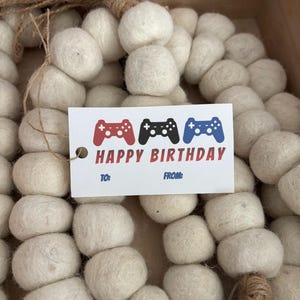 Puede incluir: Una caja de regalo llena de bolas de fieltro blanquecinas y una etiqueta que dice "Happy Birthday". La etiqueta presenta gráficos de mandos de videojuegos rojos, negros y azules. La caja está decorada con hilo.