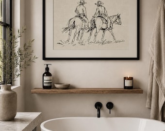 May include: Black framed artwork featuring a sketch of two cowboys on horseback. The artwork hangs above a wooden shelf with a candle, soap dispenser, and small bowl. The bathroom has a white tub and black fixtures.