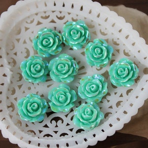 May include: Ten teal blue resin rose flower embellishments on a white lace background. The roses are all the same size and have a slightly iridescent finish.