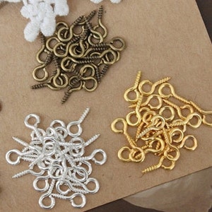 May include: Assortment of screw-in eye hooks in gold, silver, and bronze finishes. The hooks are arranged in three piles on a brown surface.