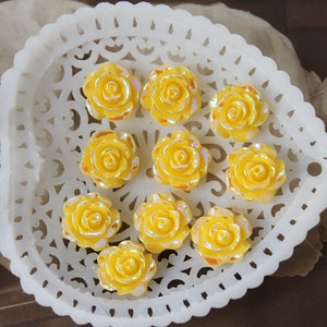 May include: A collection of ten yellow iridescent plastic rose flower embellishments displayed on a white doily.