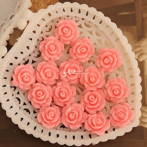 Può includere: Una collezione di 15 decorazioni di rose in resina rosa in un piatto bianco a forma di cuore. Le rose hanno un diametro di circa 2,5 cm.