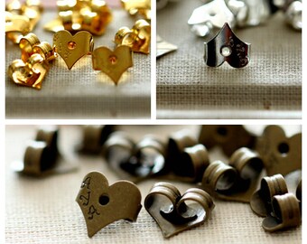 Op de afbeelding: Een assortiment van hartvormige oorbelruggen in goud, zilver en brons. Sommige ruggen hebben een klein gaatje in het midden, terwijl andere een kleine inkeping hebben. De ruggen zijn allemaal van metaal.