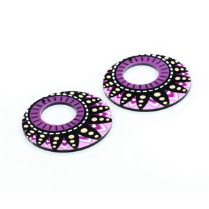 May include: A pair of round, wooden earrings with a pink, white, and black geometric design. The earrings have a large hole in the center.