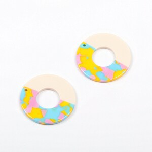 May include: Two round, white, resin earrings with a hole at the top. The bottom half of each earring is painted with a swirling pattern of yellow, pink, and blue.
