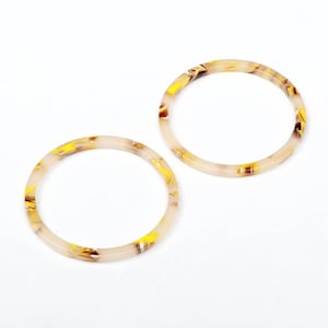 Puede incluir: Dos pendientes de aro de plástico con estampado blanco y amarillo. Los pendientes son redondos y tienen un acabado brillante.