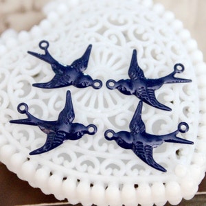Könnte beinhalten: Vier marineblaue Metall-Schwalben-Anhänger mit einer Schlaufe zum Aufhängen. Die Anhänger befinden sich auf einem weißen Hintergrund mit einem dekorativen Spitzenmuster.