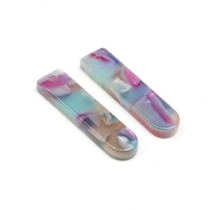 Könnte beinhalten: Zwei lange, rechteckige, irisierende Acryl-Anhänger mit einem marmorierten Muster in Blautönen, Rosa und Weiß. Die Anhänger haben oben ein Loch zum Anbringen an Schmuck oder anderen Bastelarbeiten.