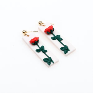 May include: A pair of white rectangular earrings with a red rose and green leaves design.