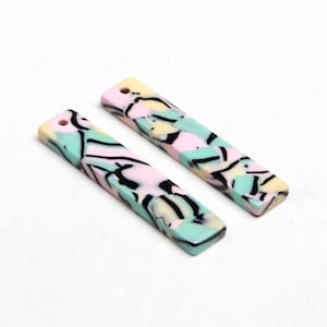 Könnte beinhalten: Zwei rechteckige, farbenfrohe Acryl-Anhänger mit einem Loch oben. Die Anhänger haben eine Pastell-Farbpalette aus Rosa, Mintgrün und Gelb mit schwarzen abstrakten Designs. Die Anhänger sind etwa 2,5 cm breit und 7,6 cm lang.