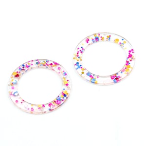 Puede incluir: Dos anillos redondos de resina transparente con confeti colorido incrustado. El confeti incluye motas rosas, azules, amarillas y doradas. Los anillos están sobre un fondo blanco, resaltando los colores vibrantes y el material transparente.
