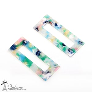 Peut inclure: Deux pinces à cheveux rectangulaires avec un motif marbré dans des tons de vert, bleu, rose et blanc. Les pinces ont une ouverture rectangulaire au centre.