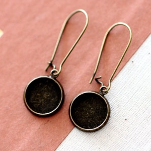 May include: A pair of antique bronze-toned earrings. Each earring features a long, curved ear wire attached to a circular bezel setting. The settings are empty, ready for customization. The earrings are displayed against a pink and cream background.