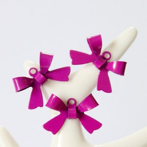 Puede incluir: Tres abalorios de metal en forma de lazo de color morado con un agujero para colgar. Los lazos son brillantes y tienen un acabado brillante.